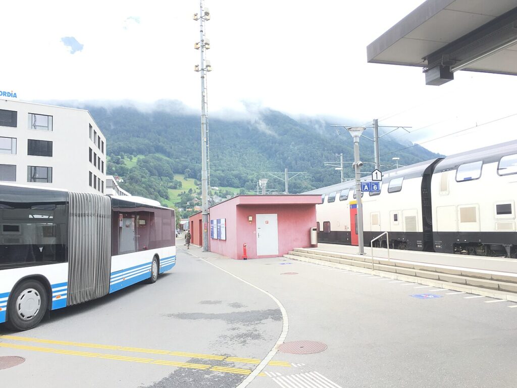 Bus und Doppelstockzug am Bahnhof Sargans mit Berghang im Hintergrund.