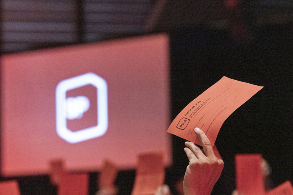 Eine Hand hält eine rote Abstimmungskarte vor einer Bühne mit SP-Logo.